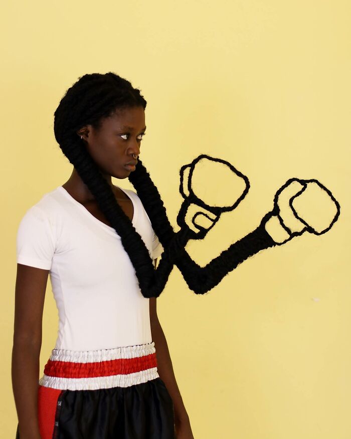 Hair sculpture resembling boxing gloves, created by an artist, worn by a person in a white shirt against a yellow background.