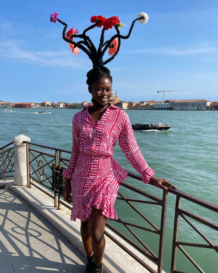 Artist with hair sculptures adorned with flowers, wearing a pink dress by the water.