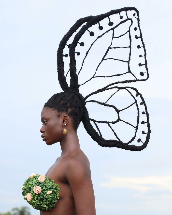 Woman with intricate butterfly hairstyle sculpture, adorned with floral decoration, set against a clear sky background.