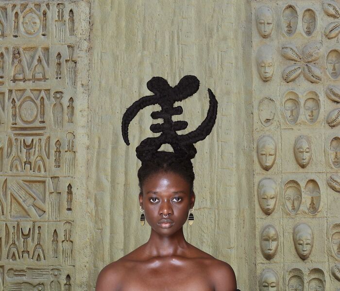 Artist with hair sculpture standing against a wall of carved faces.