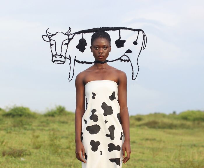Woman poses in a cow-pattern dress with a hair sculpture shaped like a cow, standing outdoors.