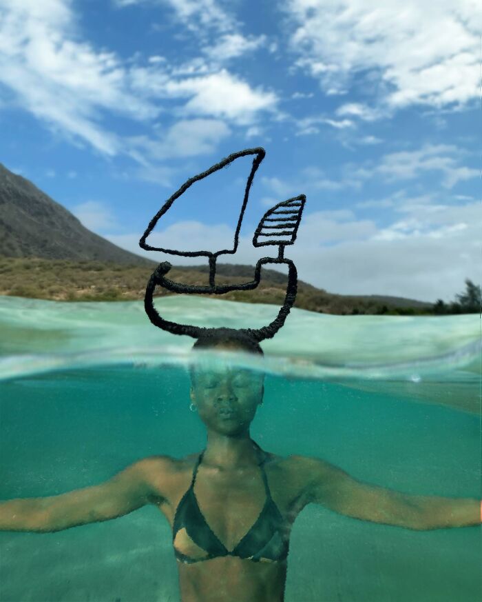 Woman with hair sculpture resembling a boat, floating in water, eyes closed, with mountain and sky in the background.