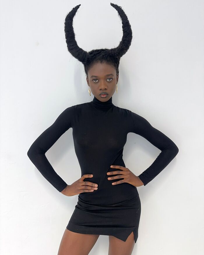 Artist with hair styled into horn-like sculpture, wearing a black outfit, against a white background.