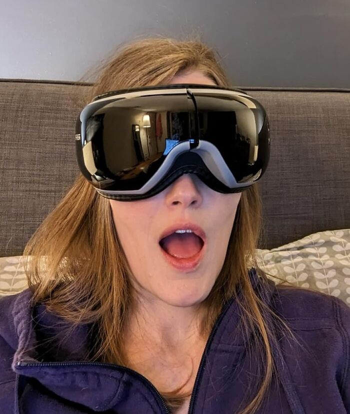 Woman wearing a futuristic eye massager sitting on a couch, reacting with surprise, a funny Mother's Day gift idea.