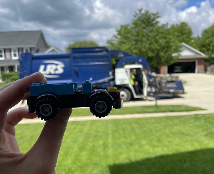 Hand holding a small toy truck in front of a real garbage truck on a bright day, capturing uplifting moments.