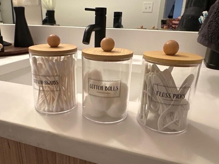 Clear bathroom storage jars with wooden lids organizing cotton swabs, cotton balls, and floss picks on a countertop.
