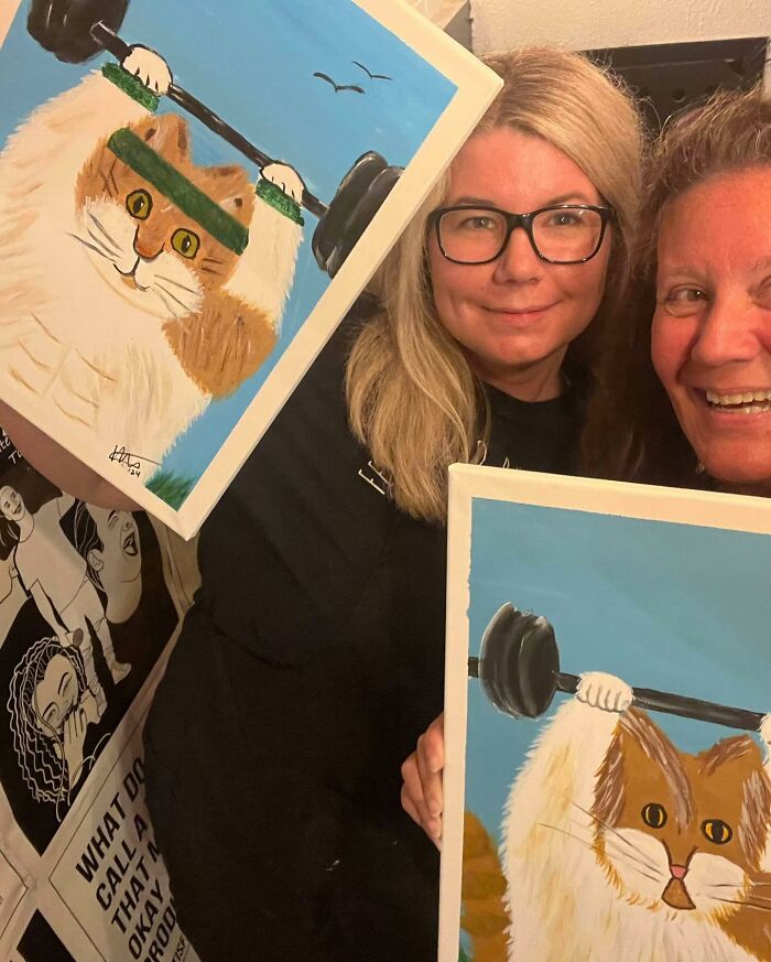 Two people smiling with paintings of a formerly overweight cat lifting weights. Two people smiling with paintings of a formerly overweight cat lifting weights.