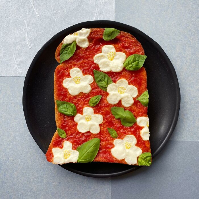Creative toast design with flower-shaped toppings and basil leaves on tomato sauce, crafted by a Japanese artist.