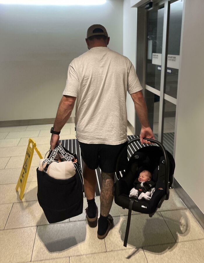 Man carrying two infants in car seats walking indoors, capturing pure and uplifting moments in daily life.