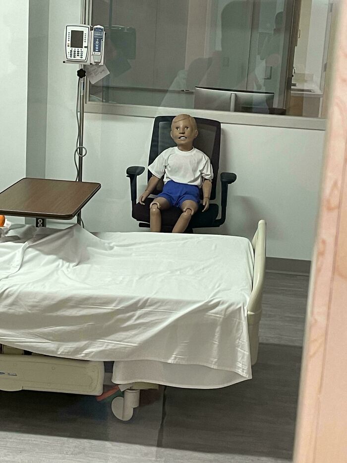 Creepy medical training dummy sitting on a chair in an empty hospital room with a bed and medical equipment nearby.