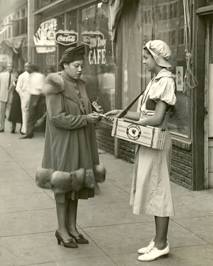African American girl selling newspapers to a woman on a city sidewalk, showcasing strength and resilience a century ago.