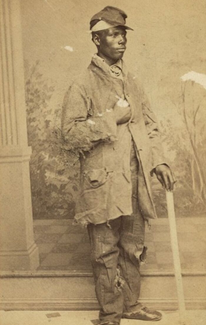 African American man dressed in worn clothing standing strong with a wooden cane showcasing strength over a century ago