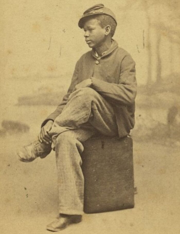 Young African American boy dressed in period clothing, sitting on a box, showcasing strength and resilience over a century ago.