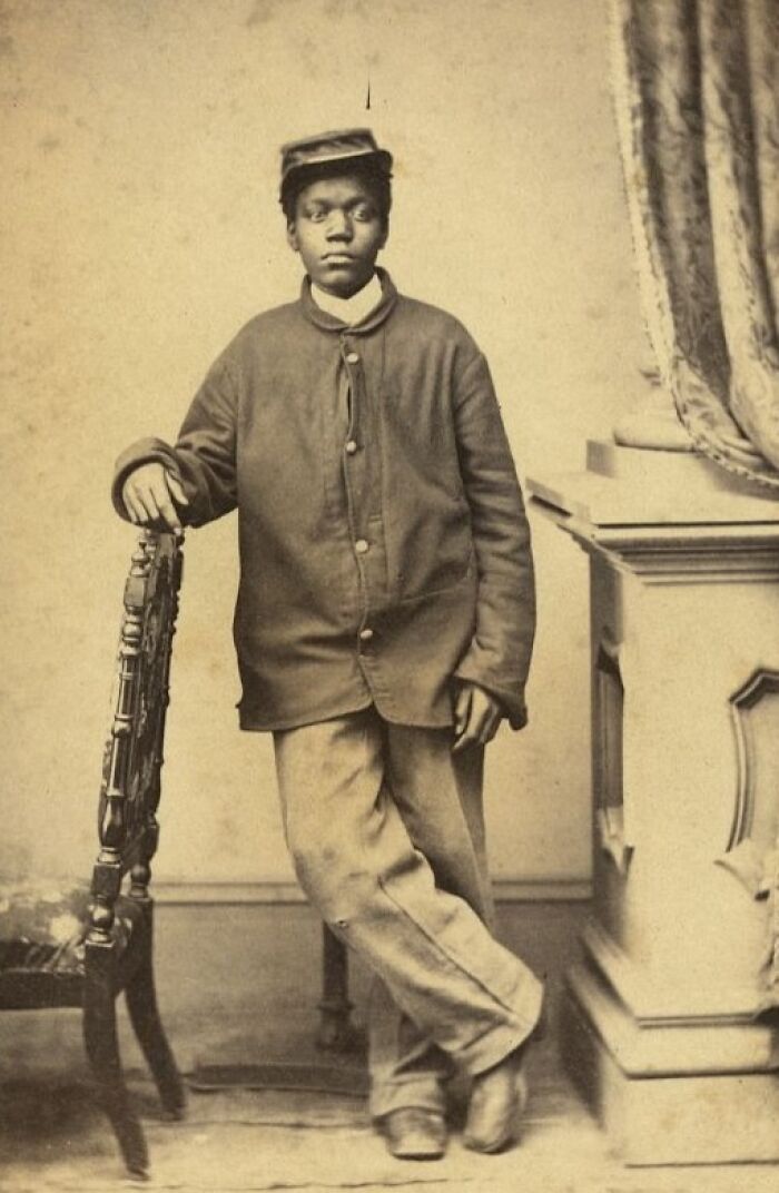 Young African American man from over a century ago in period clothing, standing confidently beside a wooden chair.