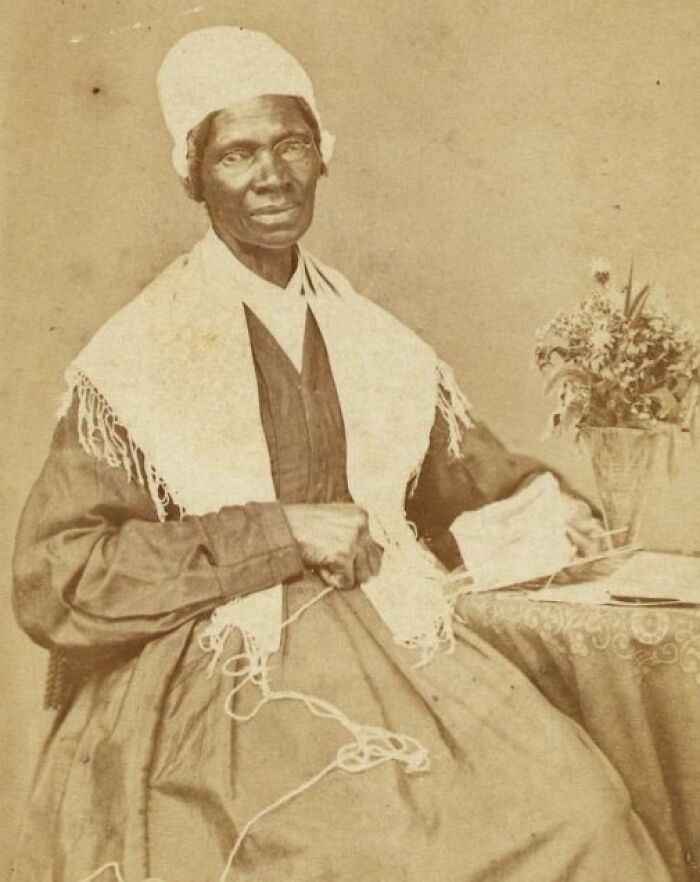African American woman from over a century ago, knitting and seated beside a table with a vase of flowers, showing strength.
