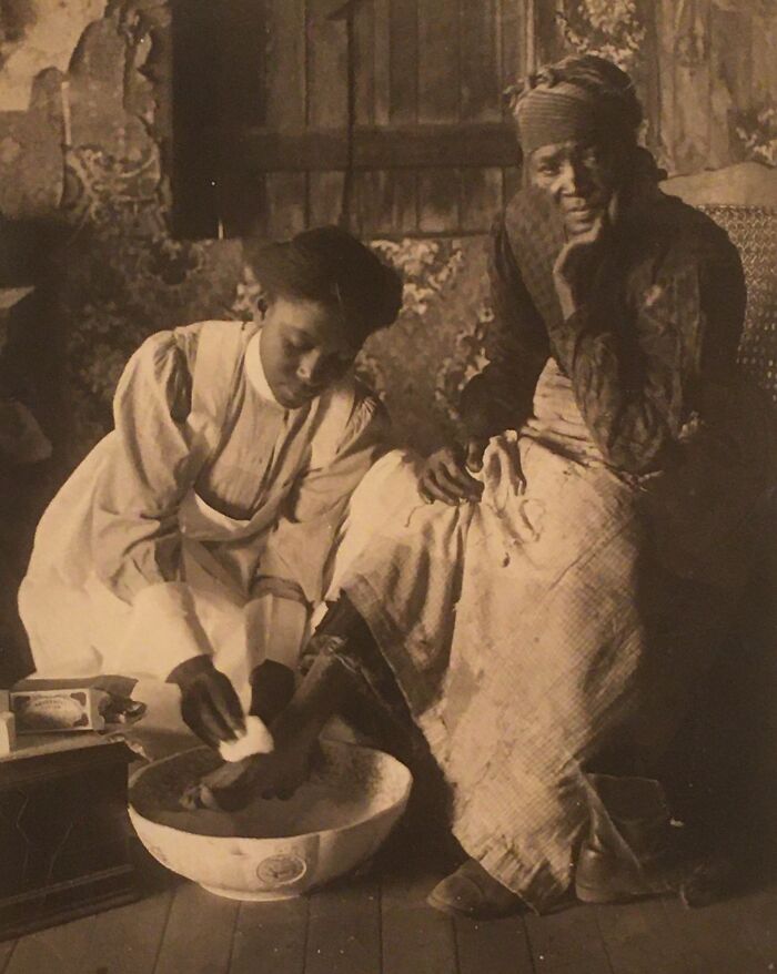 African American woman bathing elder’s feet inside a rustic room, showing strength and care over a century ago.