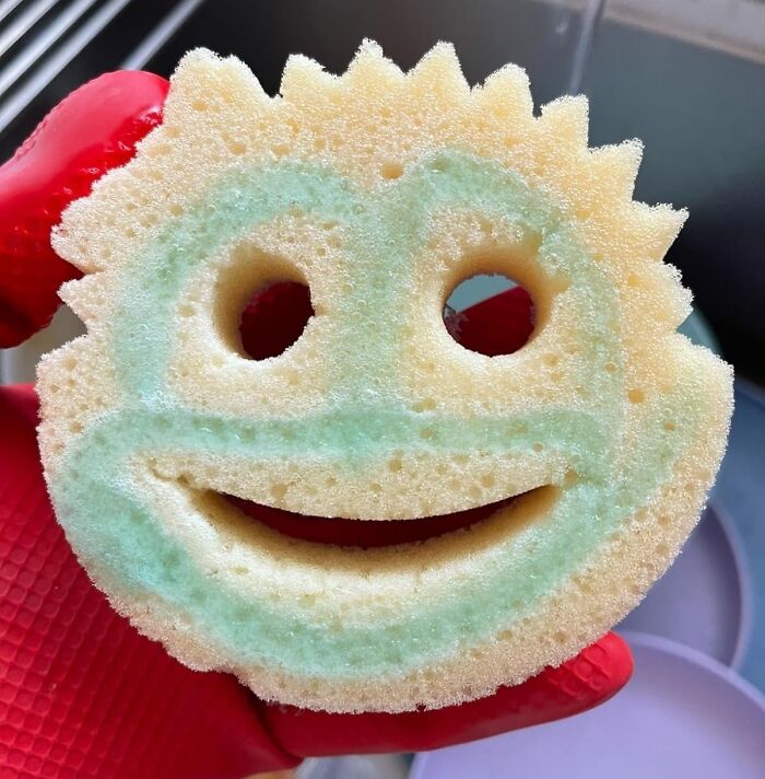 Smiling yellow kitchen sponge with cutout eyes, held by a red glove, showing a cheerful vibe for Wednesday reasons.