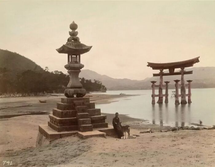 Stone lantern and traditional torii gate by the water with person and dog, depicting Japan in the 1800s.