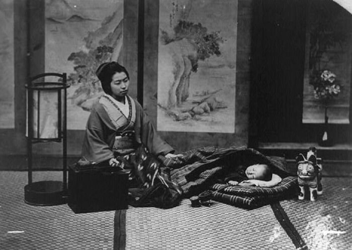 Japanese woman in traditional kimono sitting next to a sleeping child in a 1800s Japan historical indoor setting.