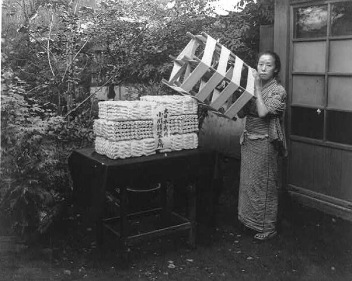 Japanese woman in traditional clothing handling wooden crates outdoors, depicting daily life in Japan in the 1800s.
