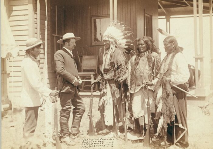 Cowboys and Native Americans in traditional attire standing and talking outside a wooden building in the 1800s.