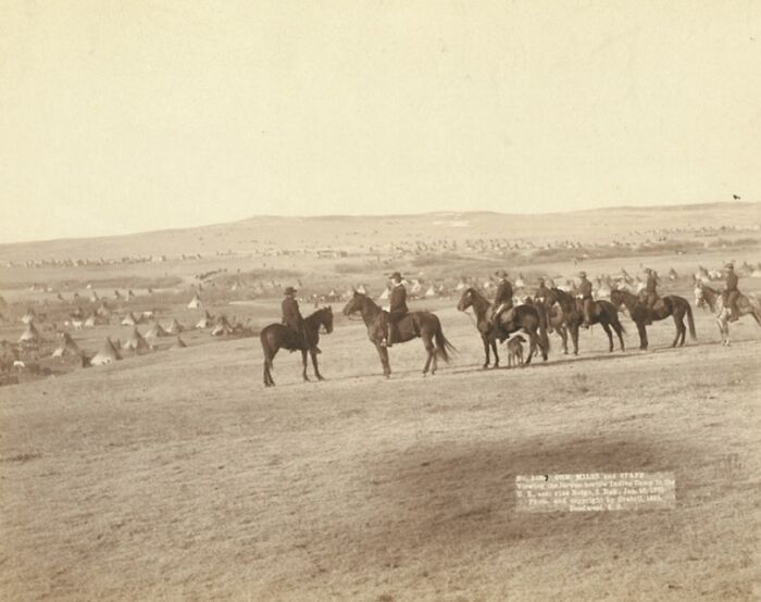 Group of cowboys and Native Americans on horseback overlooking a campsite with tipis in the 1800s landscape.
