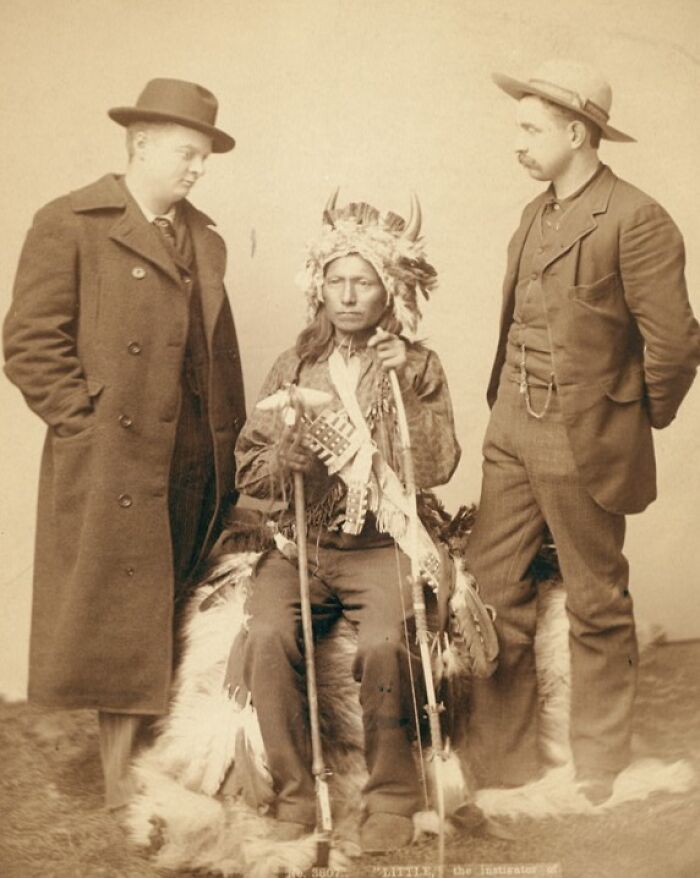 Native American in traditional attire seated between two cowboys wearing hats and late 1800s clothing.
