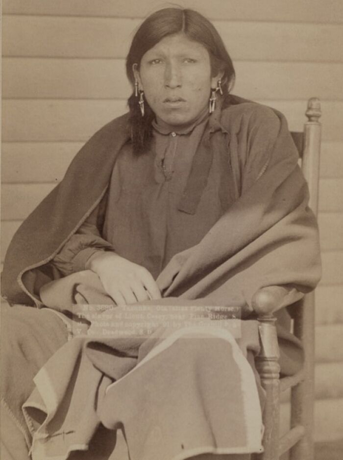 Native American man from the 1800s seated on a wooden chair, wearing traditional clothing and earrings in authentic photo.