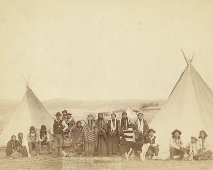 Group of Native Americans and cowboys gathered near tipis in an authentic 1800s western frontier setting.