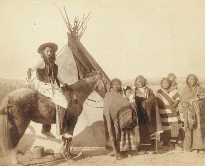 Native Americans in traditional clothing standing by a teepee with a cowboy on horseback in the 1800s West.