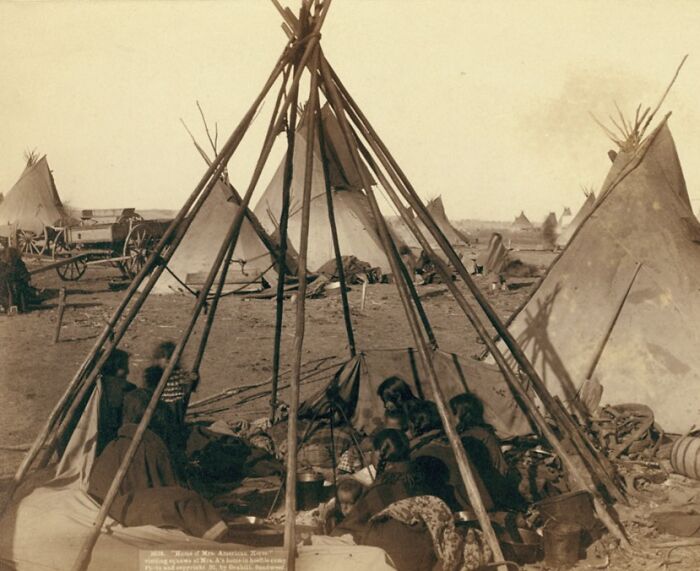 Native Americans gathered inside a traditional tipi encampment with other tipis and a wagon in the 1800s scene.