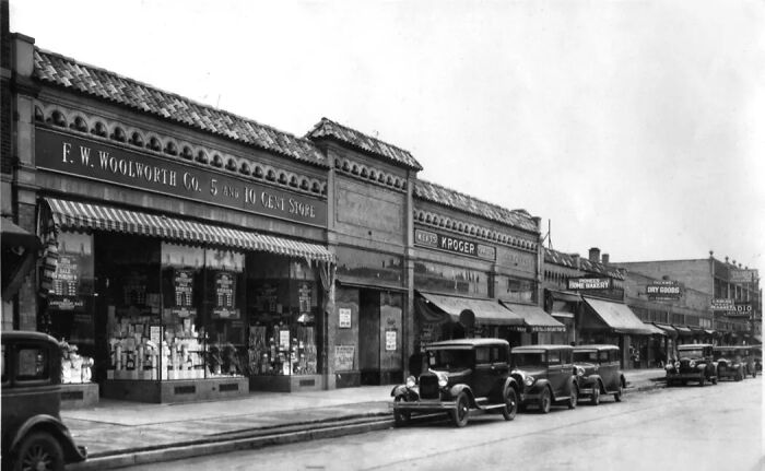 Vintage street scene showing early 20th-century American life with old stores and classic cars parked along the road.