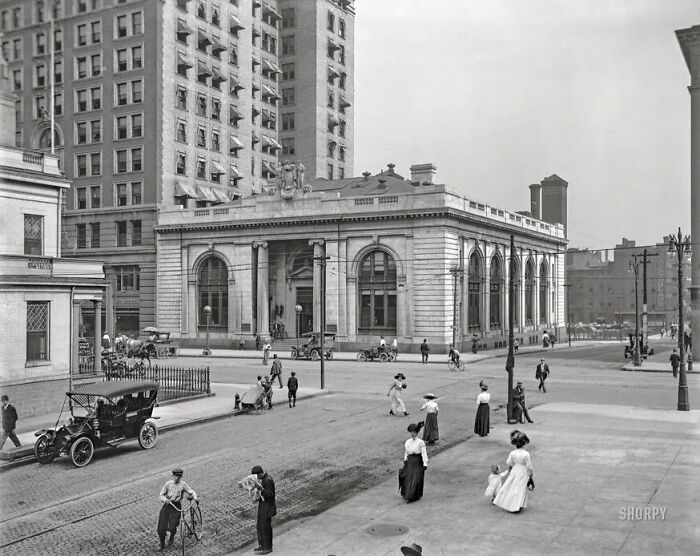Street scene in America from 100 years ago showing vintage cars, people walking, and historic buildings from the past.