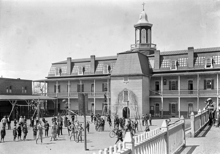 Children playing outside a large school building, sharing a glimpse of life in America from 100 years ago.