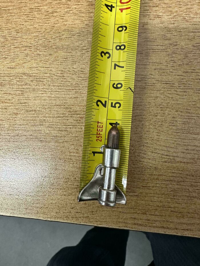 Small metal mystery object shaped like a rocket on a wooden surface next to a yellow measuring tape.