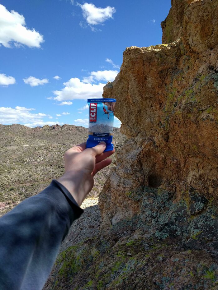 Hand holding a Clif Bar with a rocky desert landscape and blue sky in the background, an alternate angle of a natural scene.