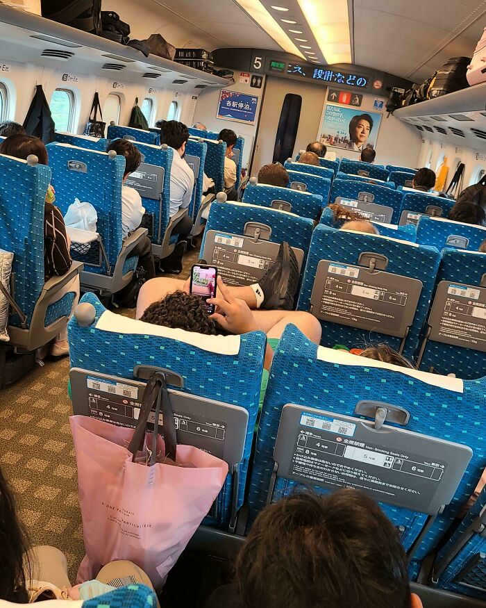Tourist lying across multiple seats indoors on a train, using a phone and blocking other passengers in crowded seating.