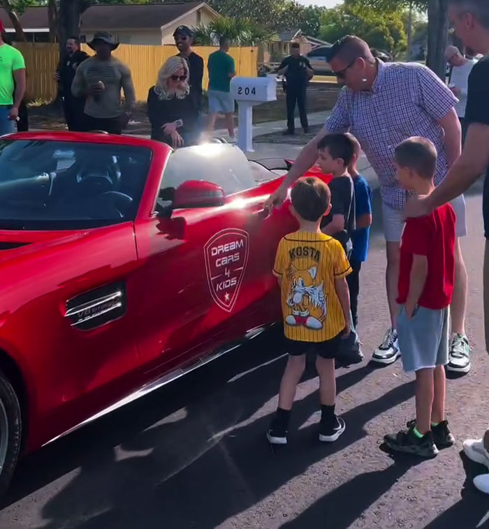 Children gathered around a red sports car with "Dream Cars 4 Kids" logo during a birthday surprise.