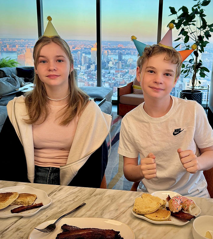 Two children in party hats sitting at a table with breakfast, enjoying a scenic view, highlighting "carbon copy" theme.