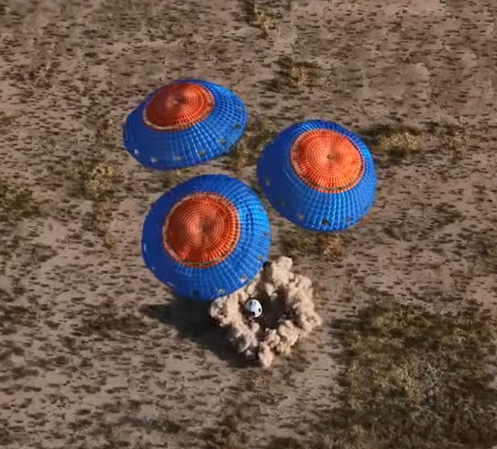Parachutes with blue and orange canopies land a capsule on a dry, grassy area, related to Blue Origin flight.