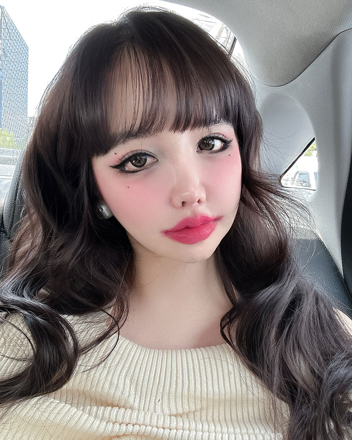 K-Pop star posing in a car, with long hair and glamorous makeup highlighting drastic transformation.
