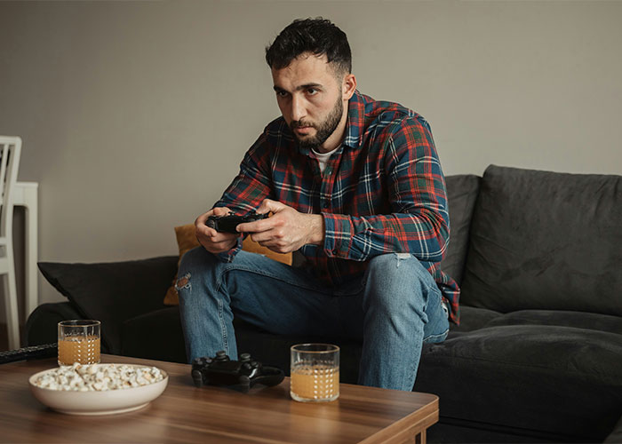 Man playing video games on a couch, flannel shirt, focused, with drinks and popcorn on the table, illustrating movie unrealisticness.