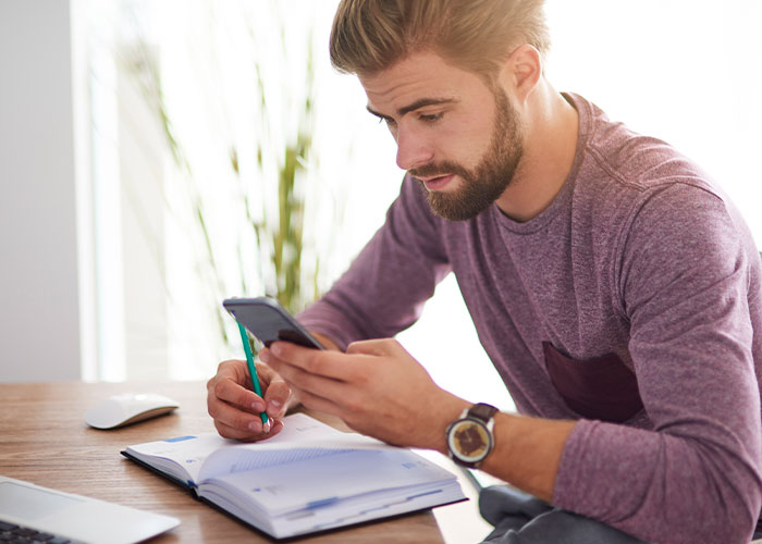 Man using phone and writing in planner at desk, exploring cheat code ideas for marriage success and tips.