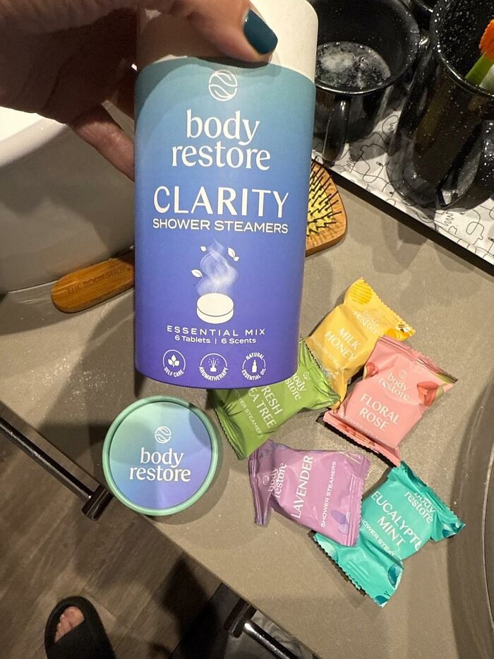 Hand holding Body Restore Clarity shower steamers container with multiple scent packets on bathroom counter.