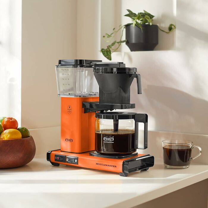 Orange Moccamaster coffee maker on a white counter, brewing coffee, surrounded by fruit and plants, lasting a lifetime.