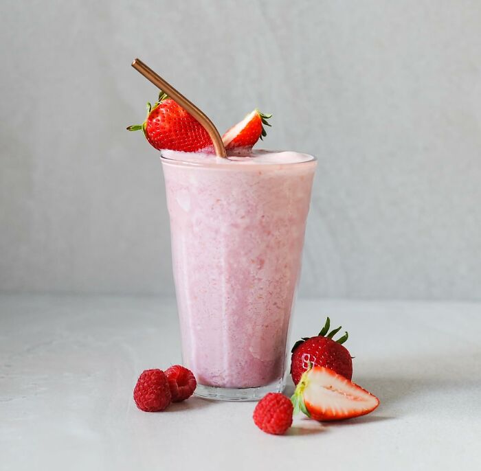 Strawberry smoothie in a glass with a metal straw, surrounded by fresh strawberries and raspberries on a light background.