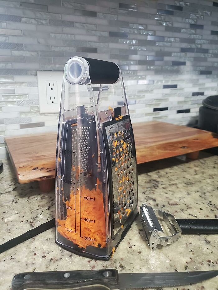 Multi-sided kitchen grater with shredded carrots inside and a vegetable peeler on a granite countertop, time savers concept.