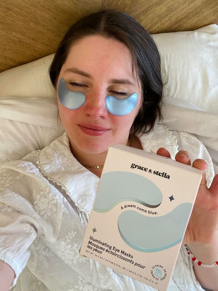 Woman with eye masks holding a box of Amazon's most-wished-for illuminating eye masks lying on a bed.