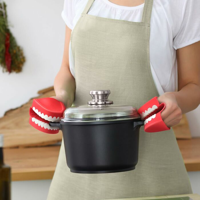 Person wearing apron holding pot with quirky teeth-shaped mitts; genius kitchen find.
