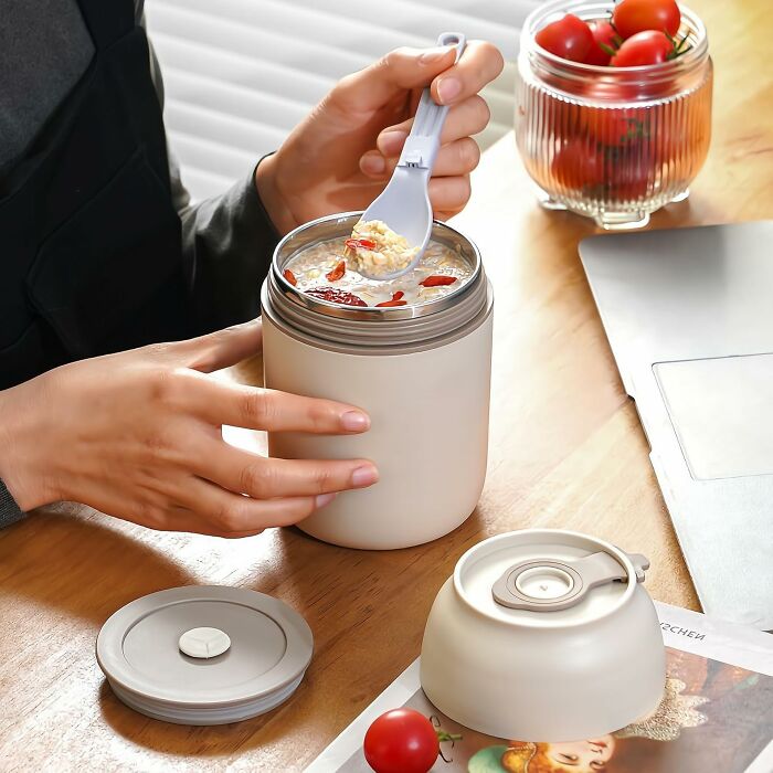 Hand holding a spoon scooping food from a beige insulated container, with cherry tomatoes and a laptop on a wooden table.